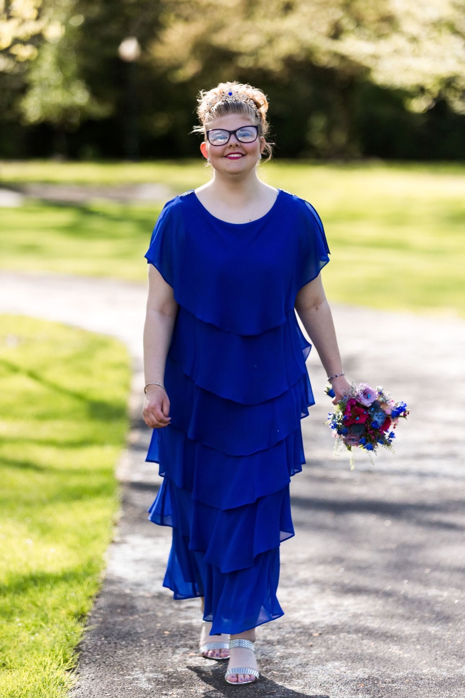 Elegant blue tiered dress with silver sandals and colorful floral bouquet on a sunny garden path.