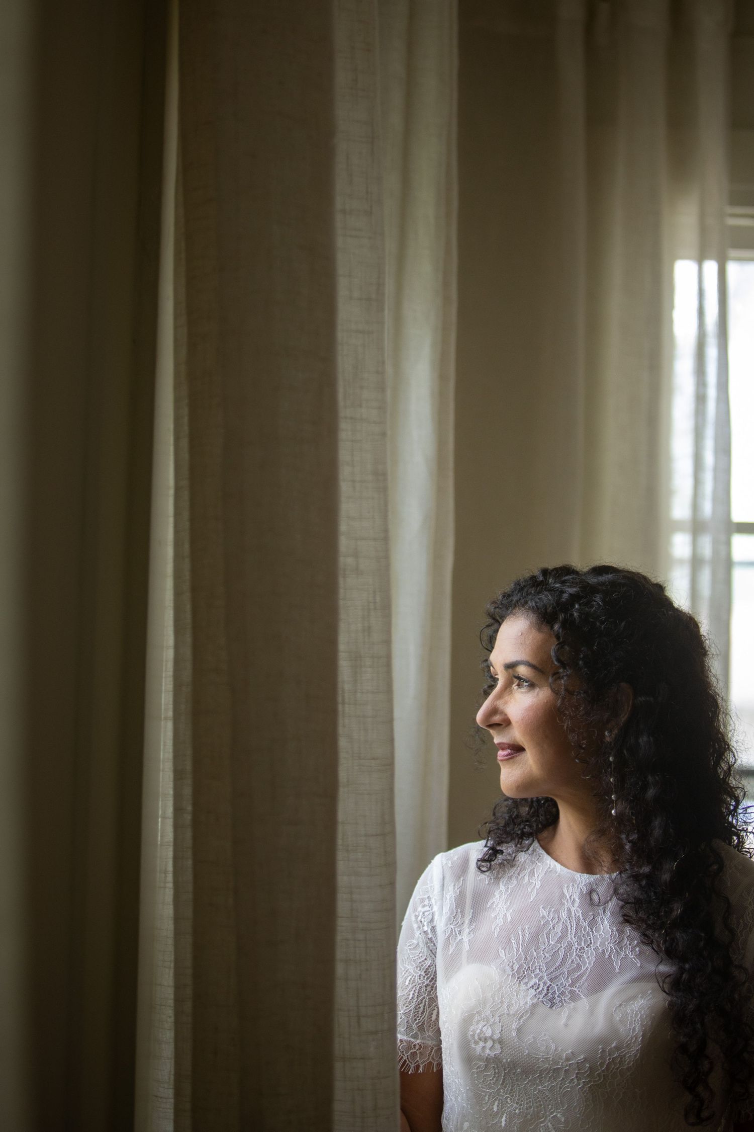 Side profile of bride in white lace dress gazing through sheer curtains in natural light.