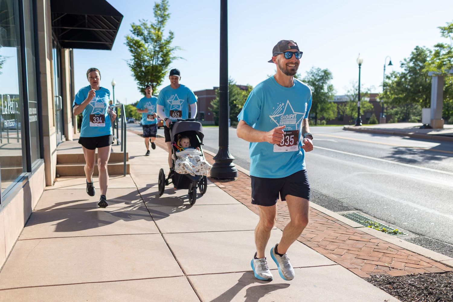 Runners in light blue shirts participate in an outdoor racing event on a city sidewalk.