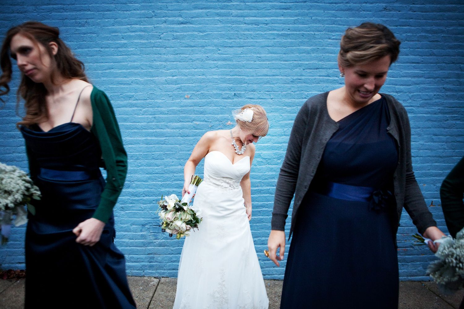 Wedding party stands against vibrant blue wall while bride laughs in white gown alongside bridesmaids in navy dresses.