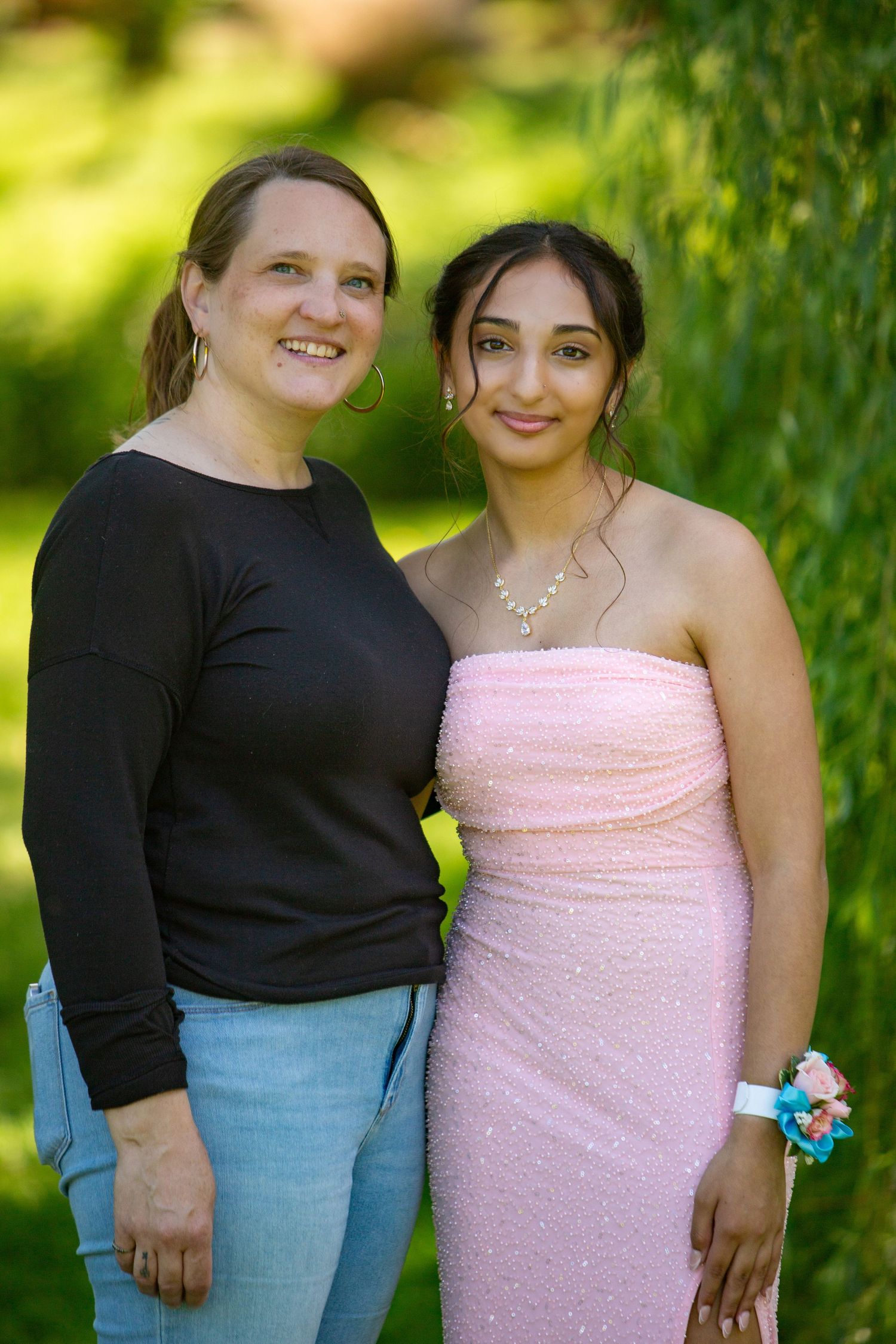 Two people stand together outdoors, one wearing a black sweater and jeans and one in a sparkly pink formal dress.