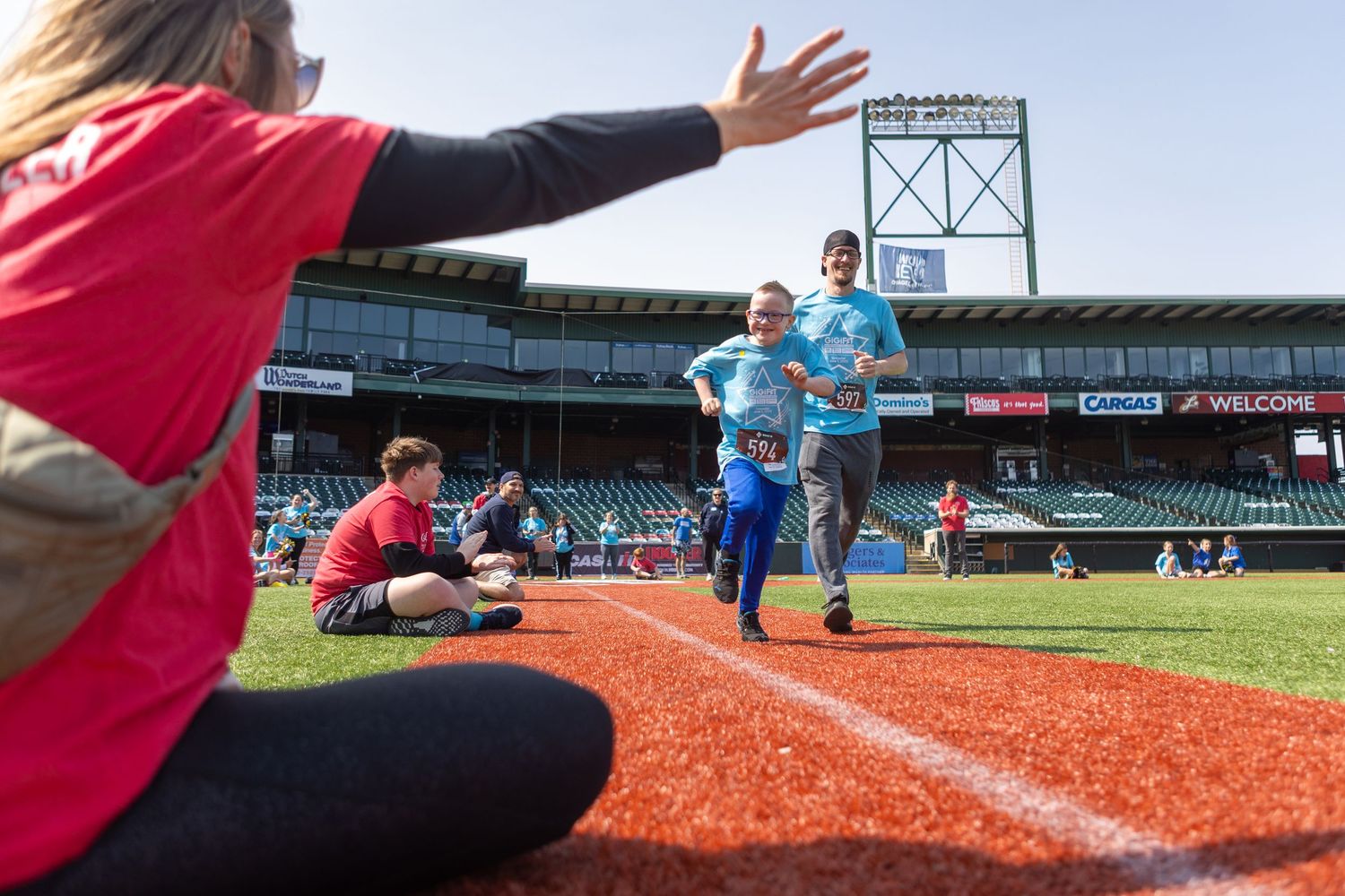 Participants run along the warning track at a baseball stadium while volunteers encourage them.