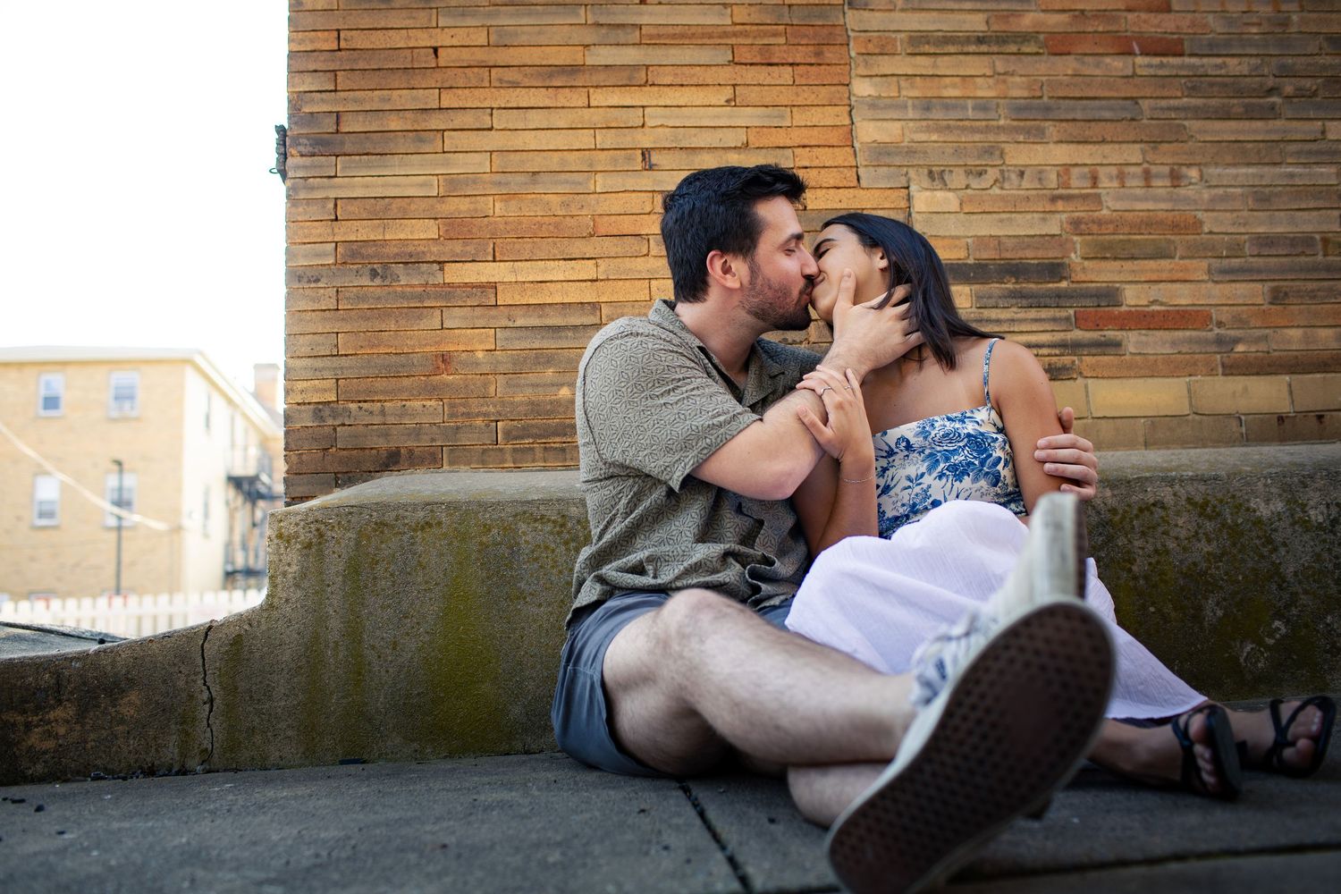 A romantic couple sits together against a brick wall on a sunny afternoon sharing an intimate moment.