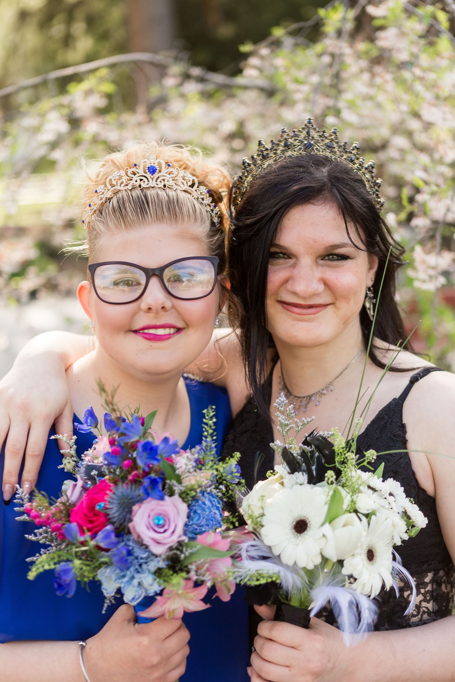 Two friends at a formal event holding colorful bouquets of flowers, one in blue and one in black, both wearing tiaras.