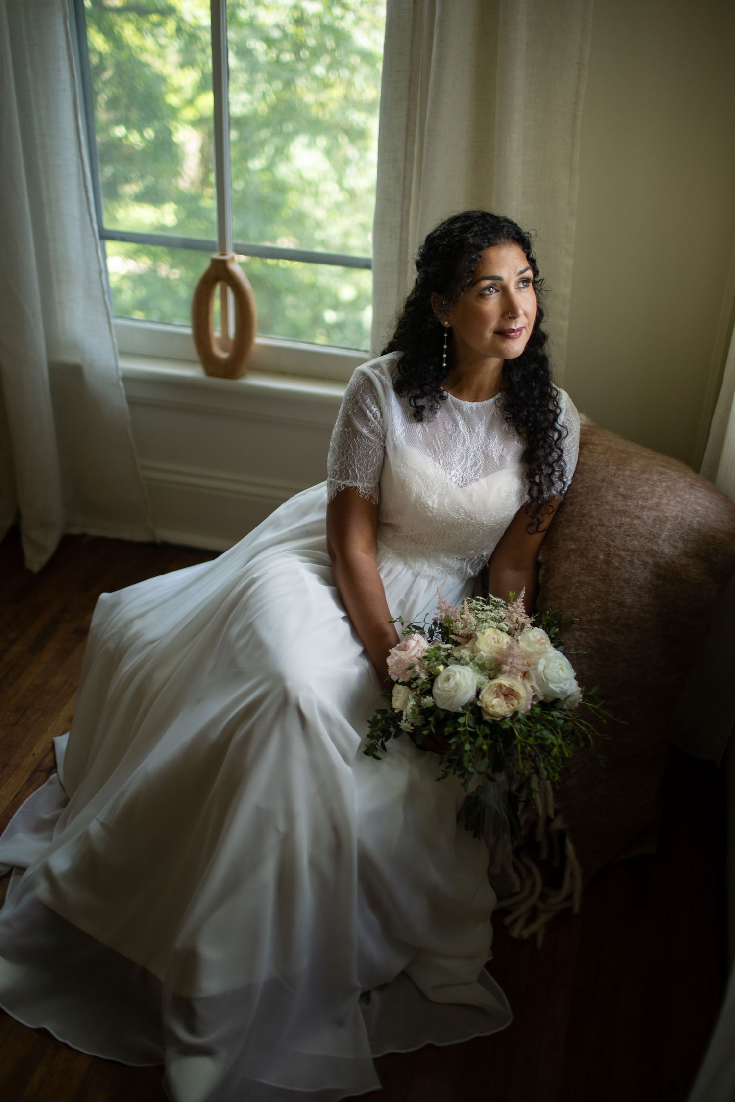 A bride in a classic white lace gown sits with her bouquet near a sunlit window.