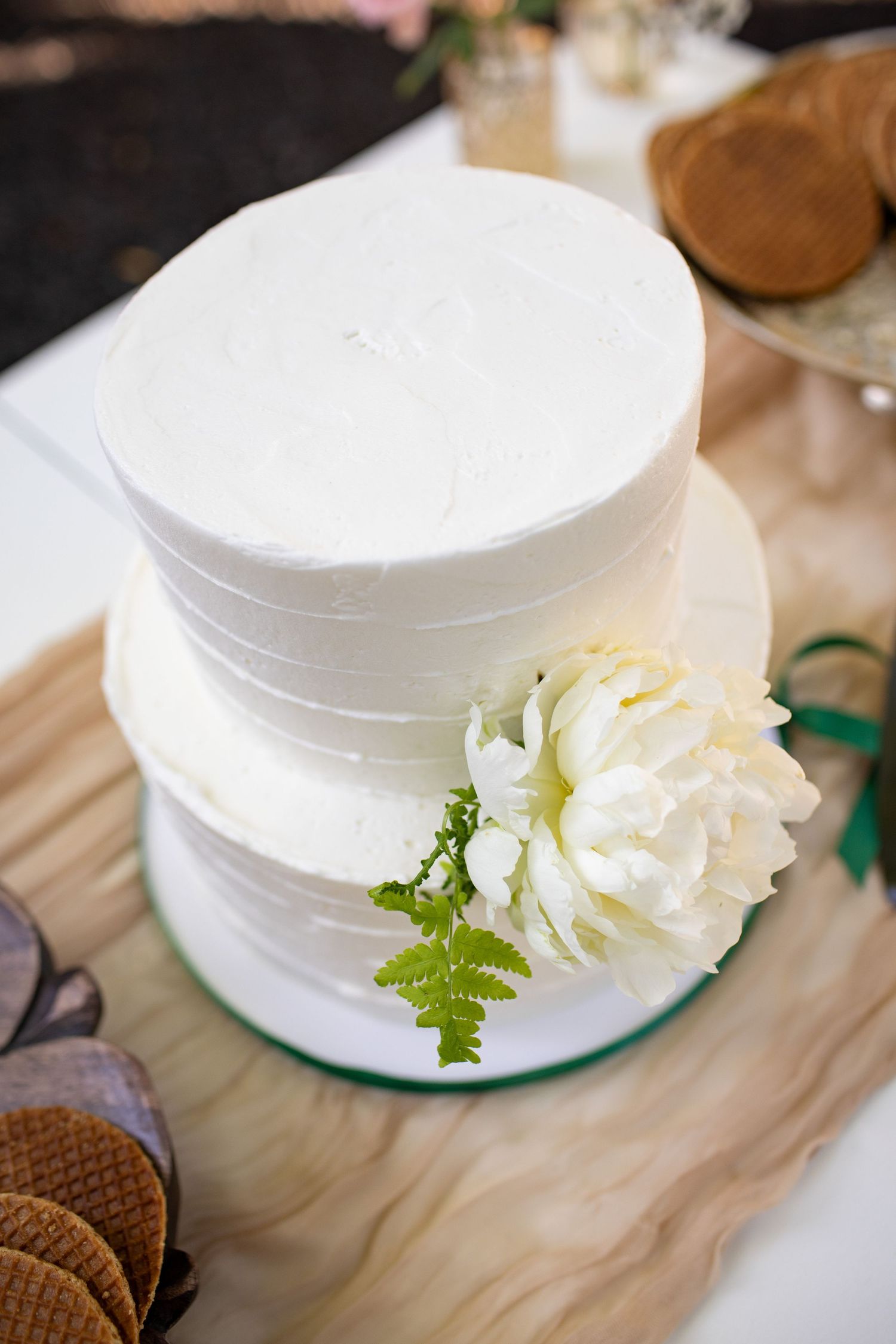 Simple two-tiered white wedding cake decorated with a fresh white peony flower and fern leaf on wooden surface.