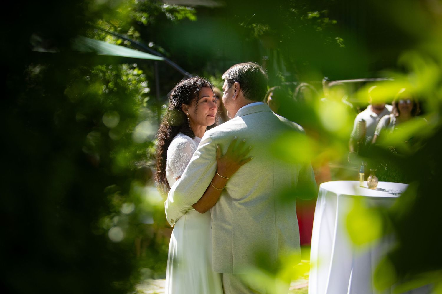 Two people in white wedding attire share a romantic kiss in a lush garden setting with soft natural lighting.