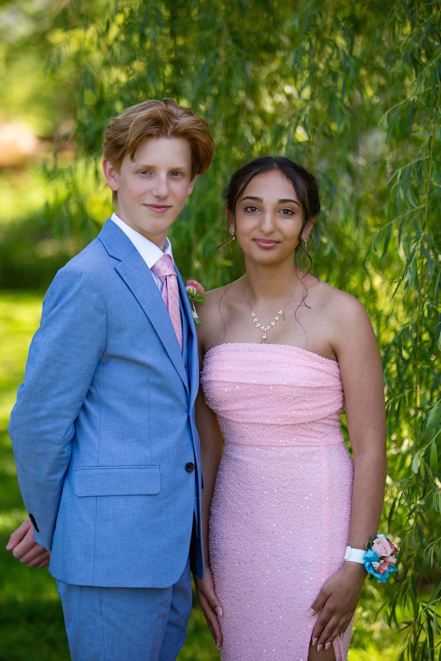 A couple in formal attire poses together under a willow tree, wearing a light blue suit and pink dress.