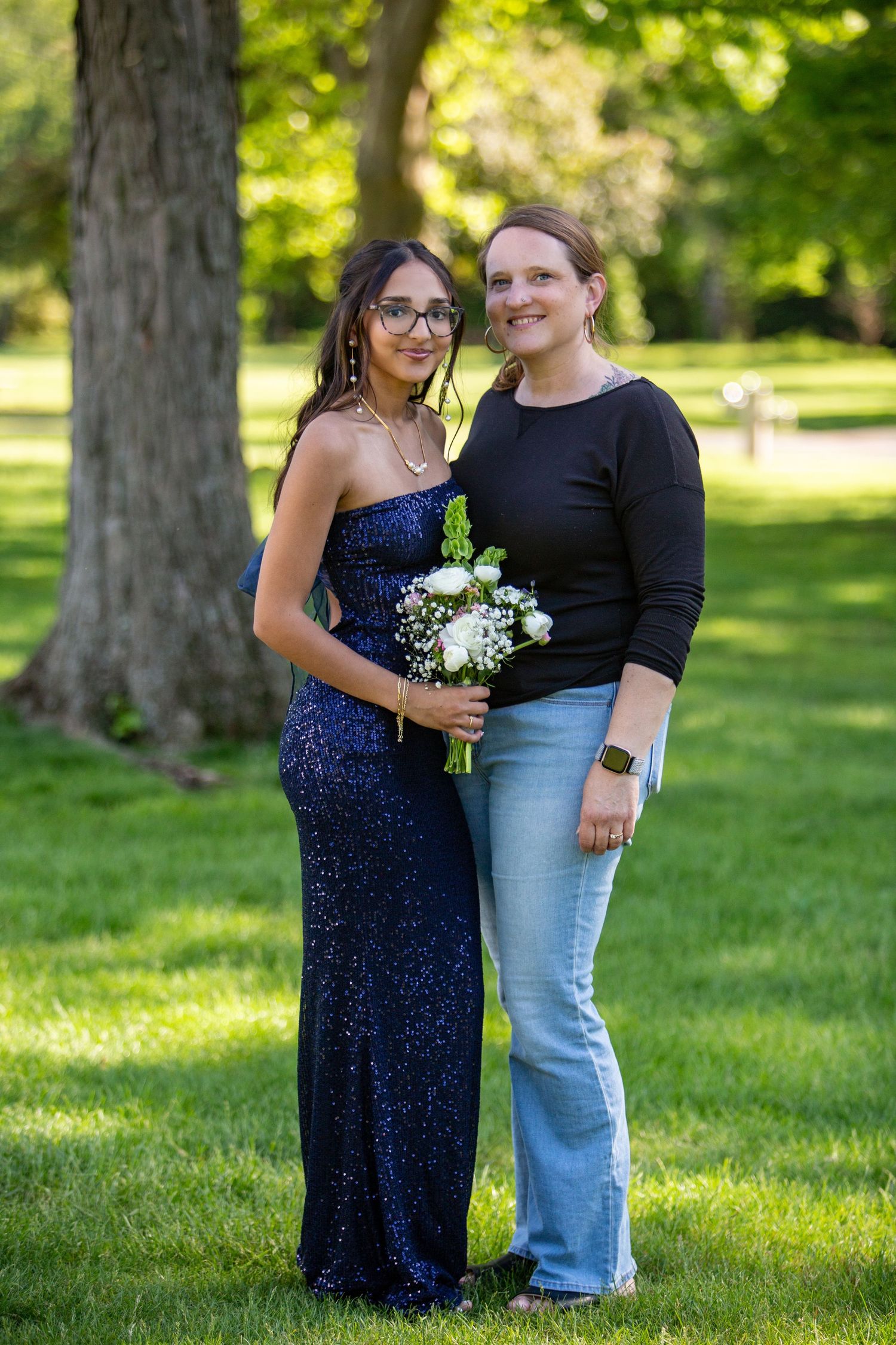 Two people stand together outdoors in a park setting, one wearing a sparkly blue gown holding white flowers.