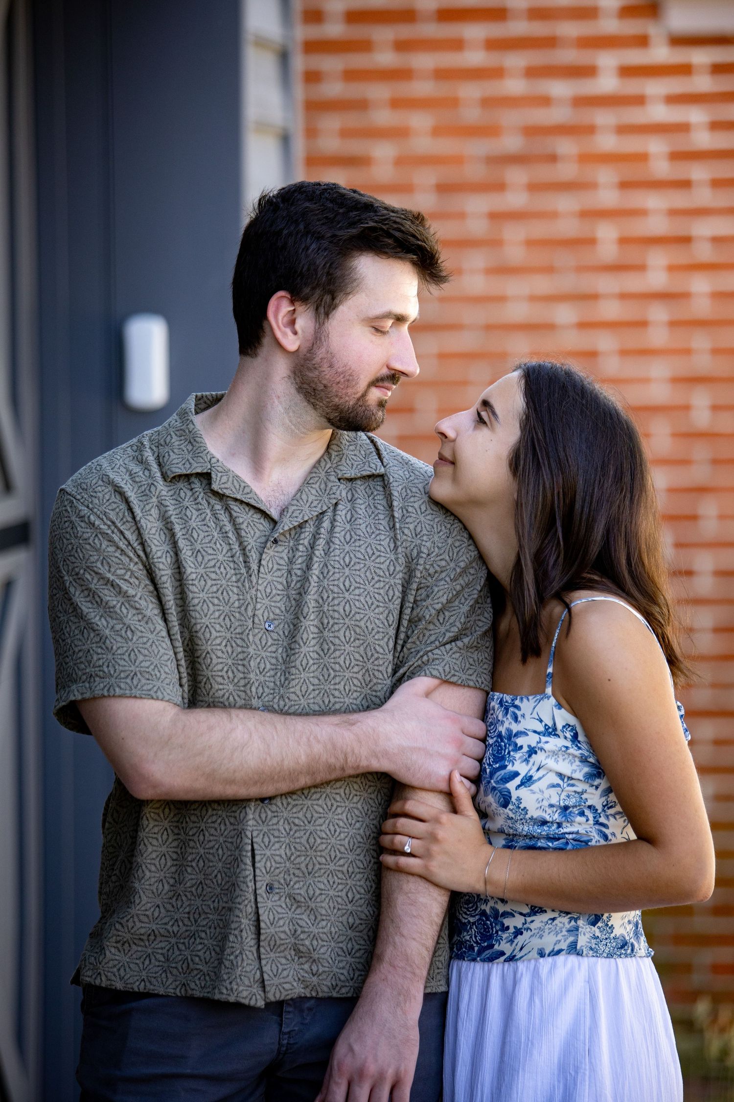 Romantic couple embraces outdoors against brick wall, with woman in floral dress and man in polo shirt.