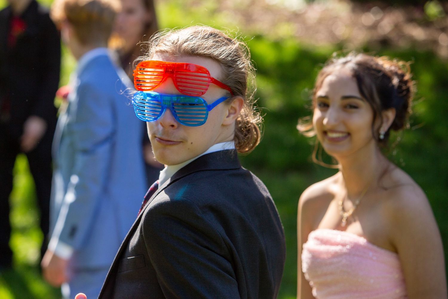 A person wearing a tuxedo and bright red sunglasses poses outdoors with someone in a pink dress.
