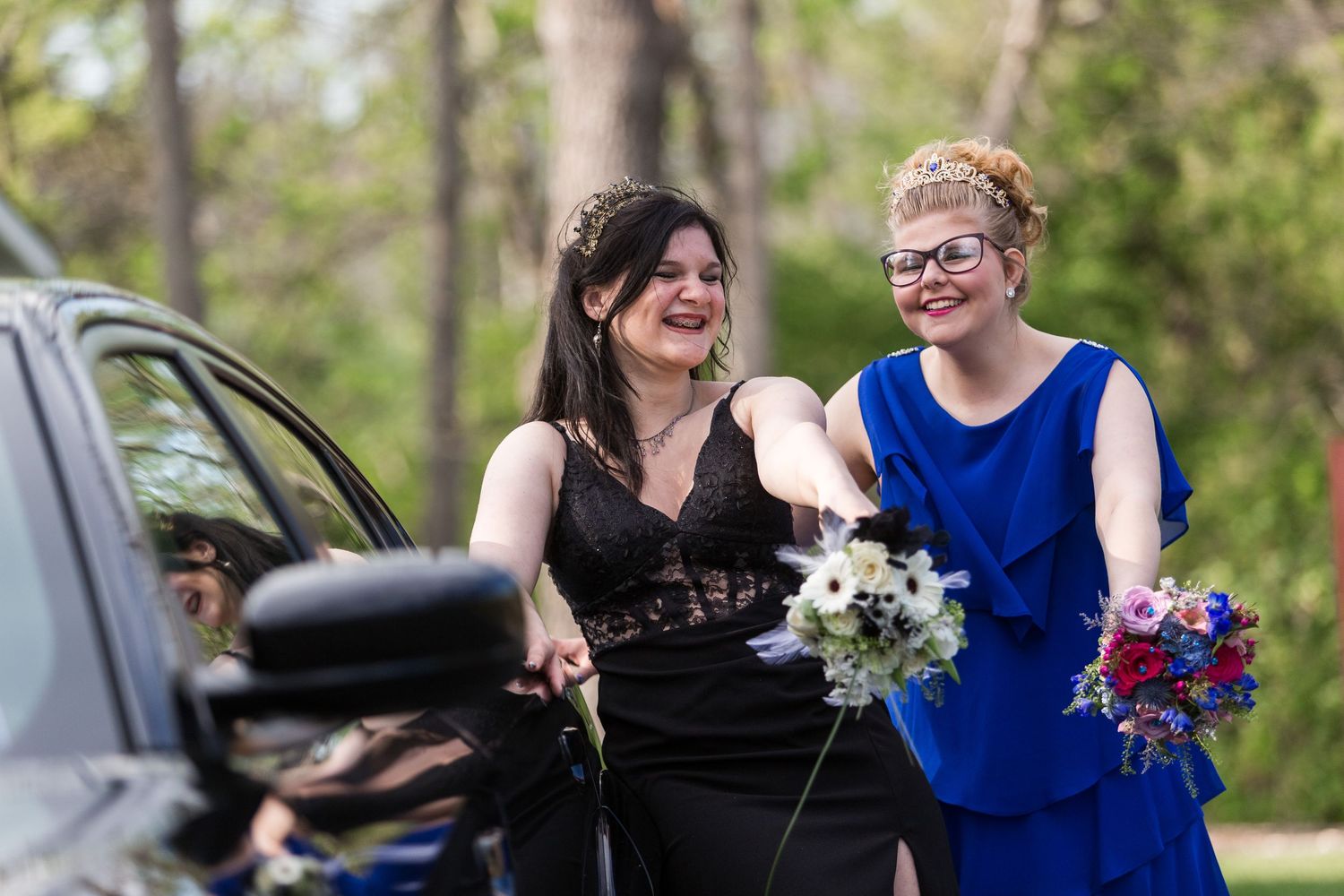 Two prom friends in black and blue dresses smile while holding colorful bouquets next to a car.