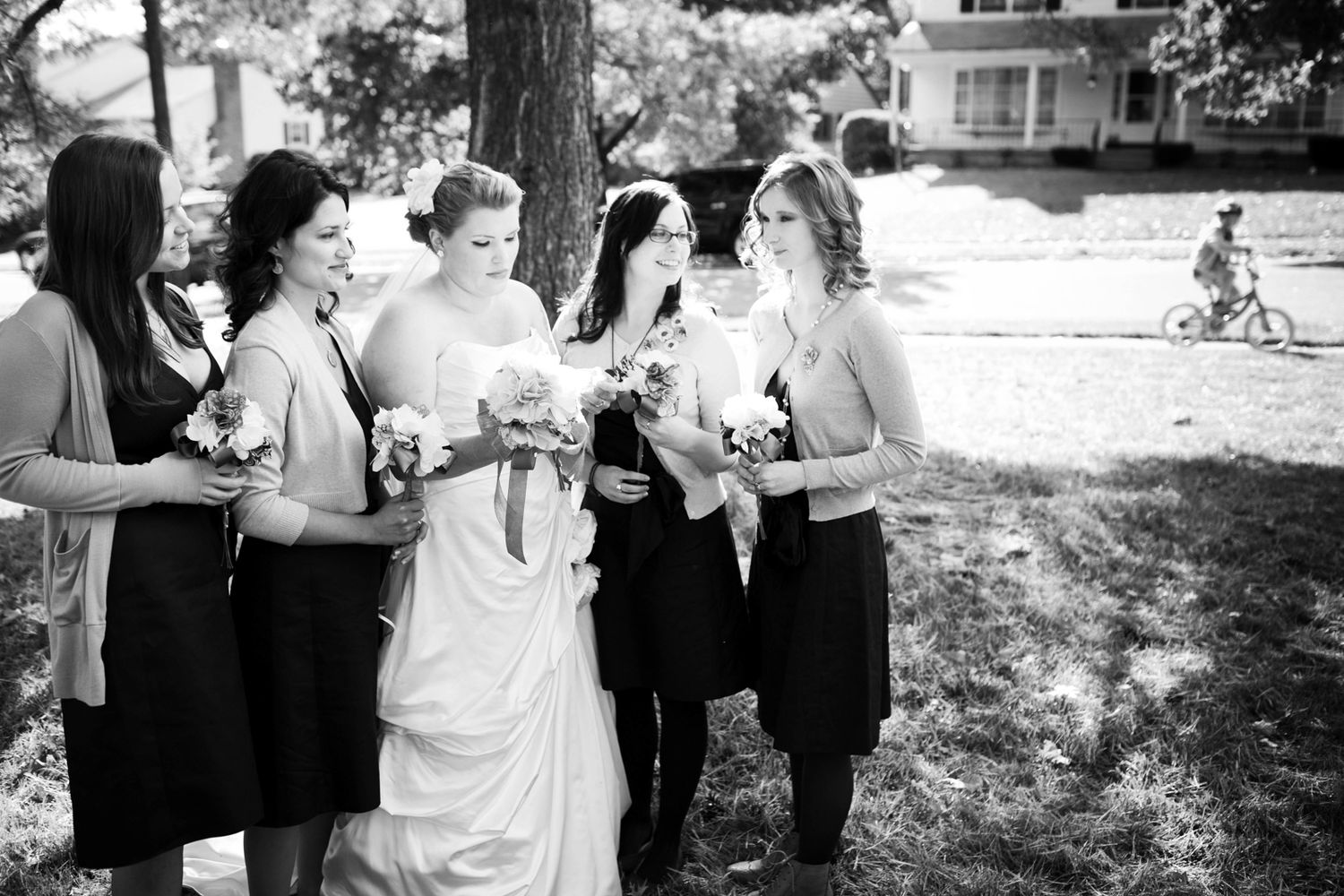 Bride and bridesmaids in black dresses gather outdoors holding bouquets during wedding celebration.