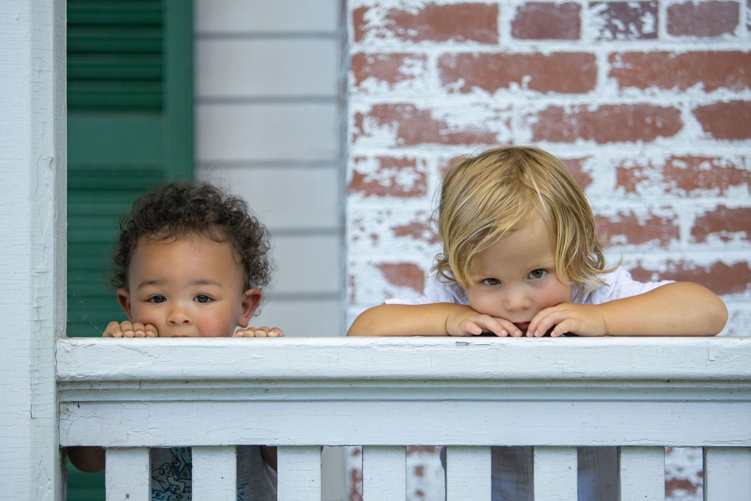 Two playful children peek over a white porch railing in front of a brick wall with green shutters in the background.