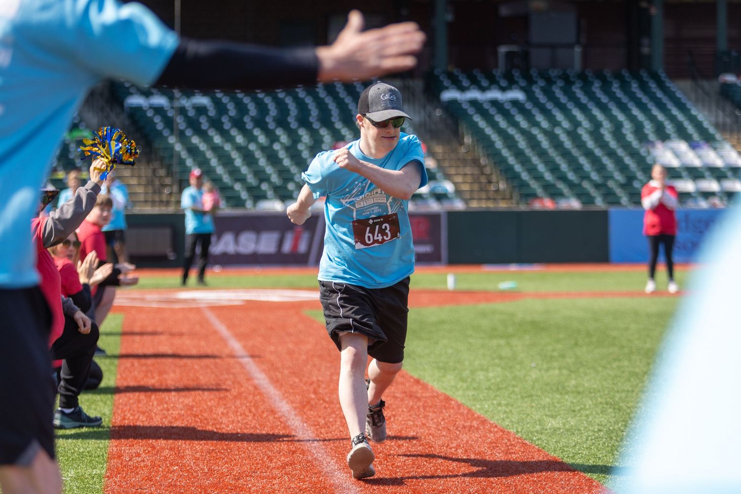 Runner in light blue shirt and cap running along baseball field warning track.