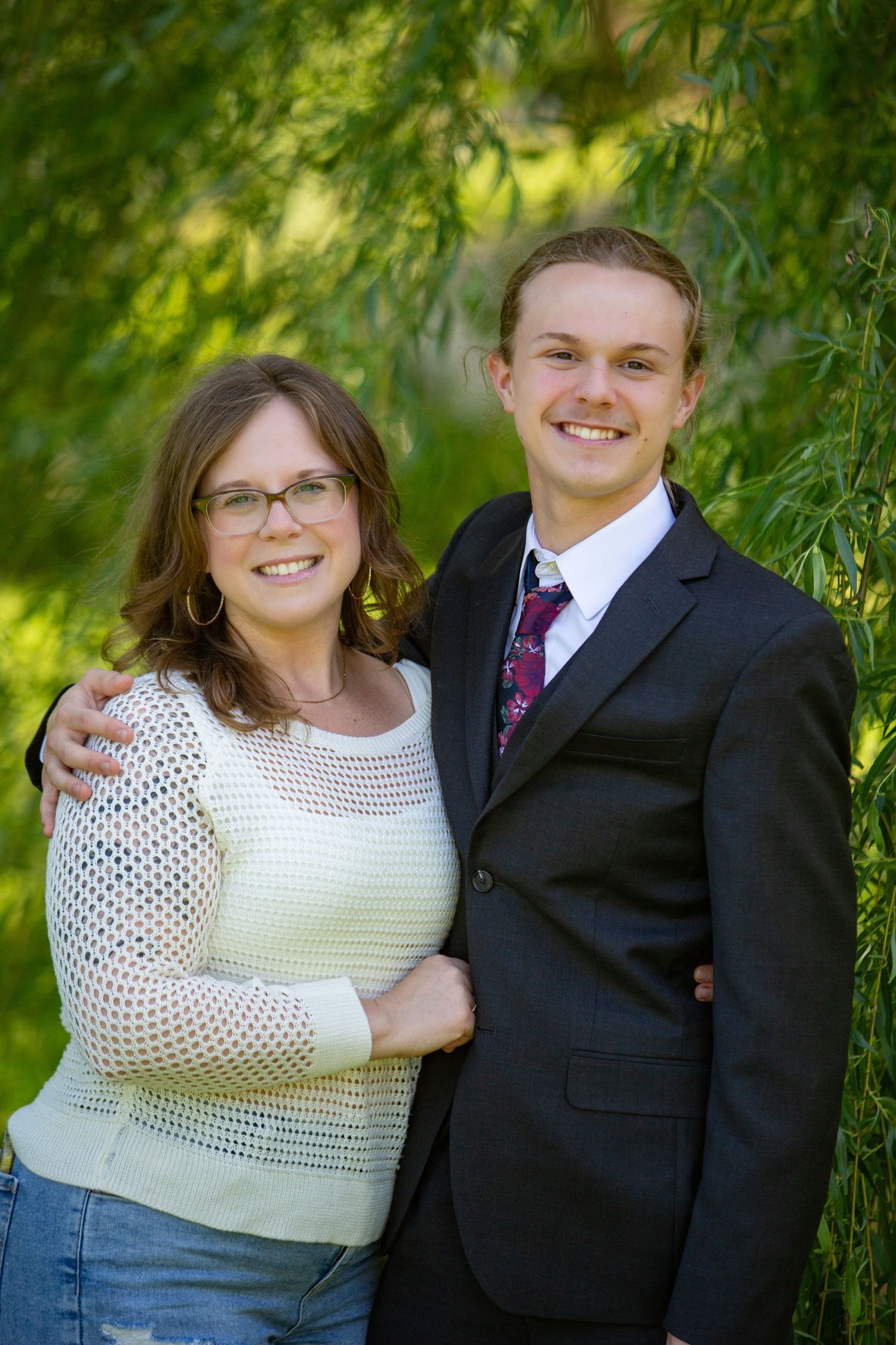 A couple poses together outdoors, with a leafy green backdrop, wearing a white sweater and dark suit with floral tie.