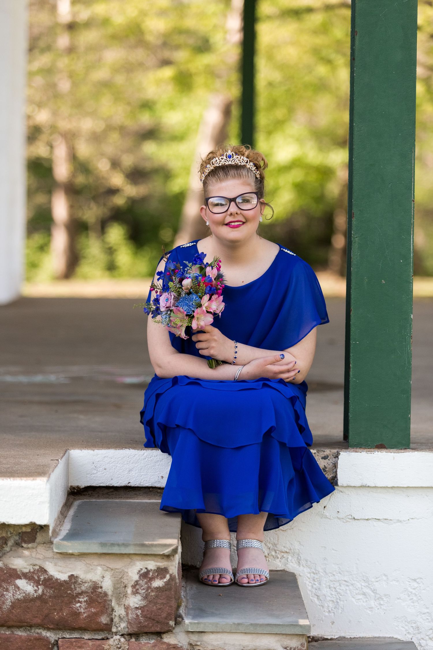 A cheerful figure in a royal blue layered dress with glasses sits on stone steps outdoors wearing silver sandals.
