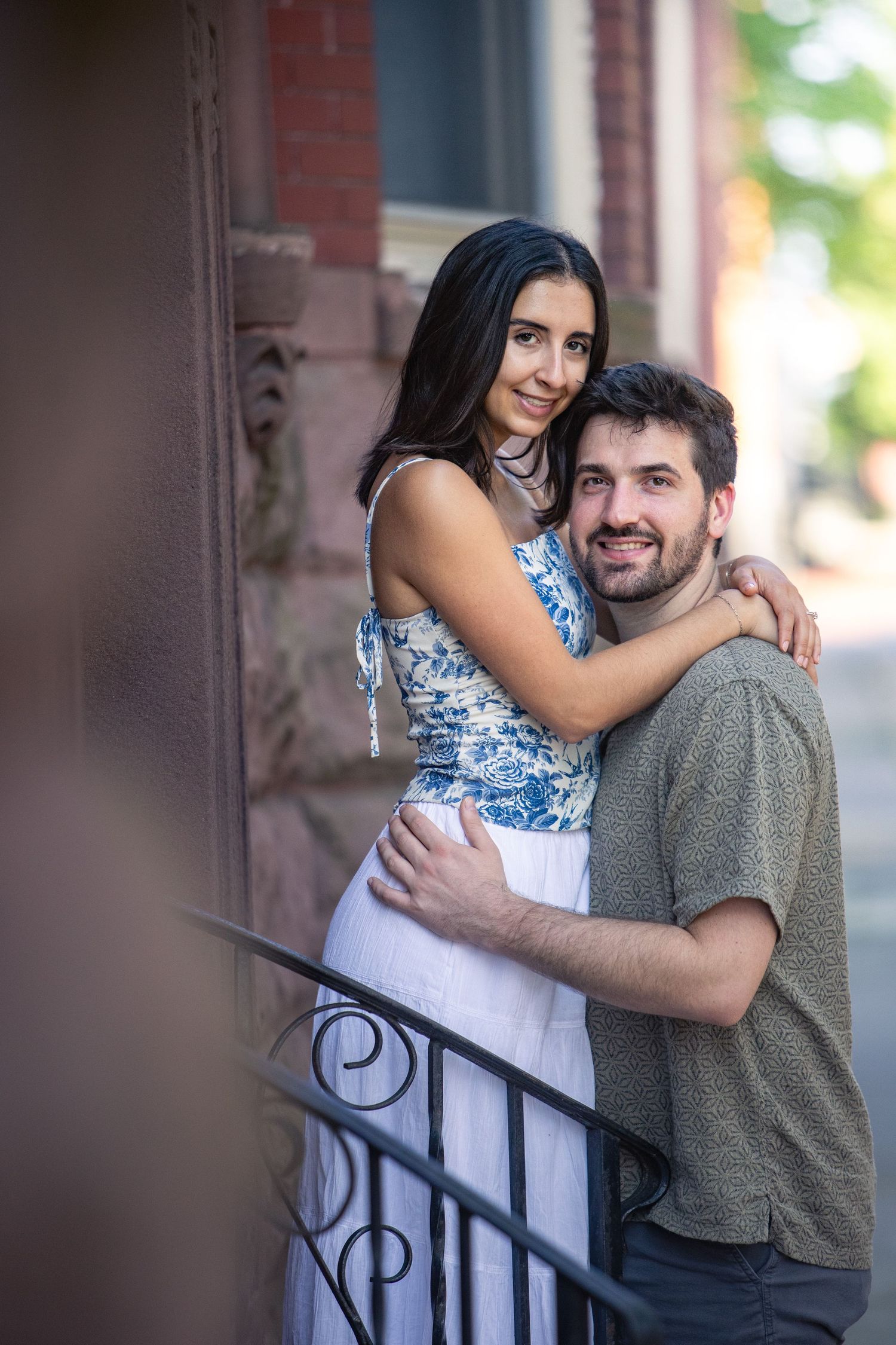 A couple embraces on a city sidewalk next to decorative iron railings and brick buildings.