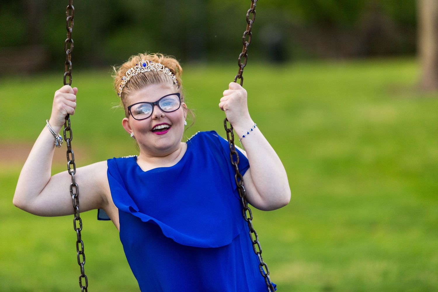 A happy person wearing glasses and a bright blue shirt enjoys swinging at a playground on a sunny day.