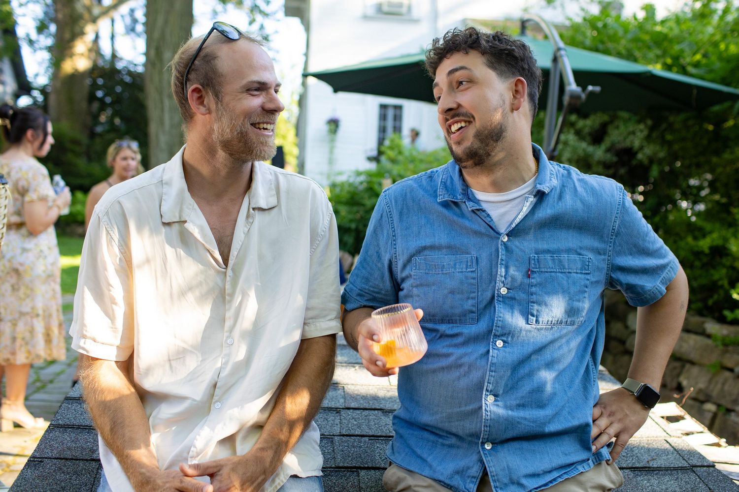Two men in casual summer attire share a laugh while enjoying drinks at an outdoor gathering.