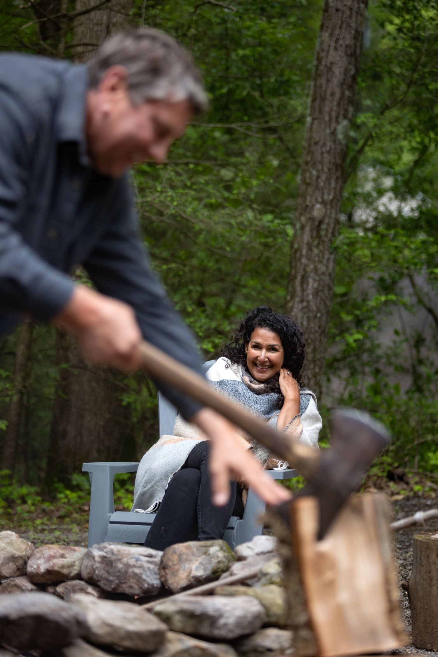 Two people sitting by a fire pit in a wooded area while one person chops wood in the foreground.