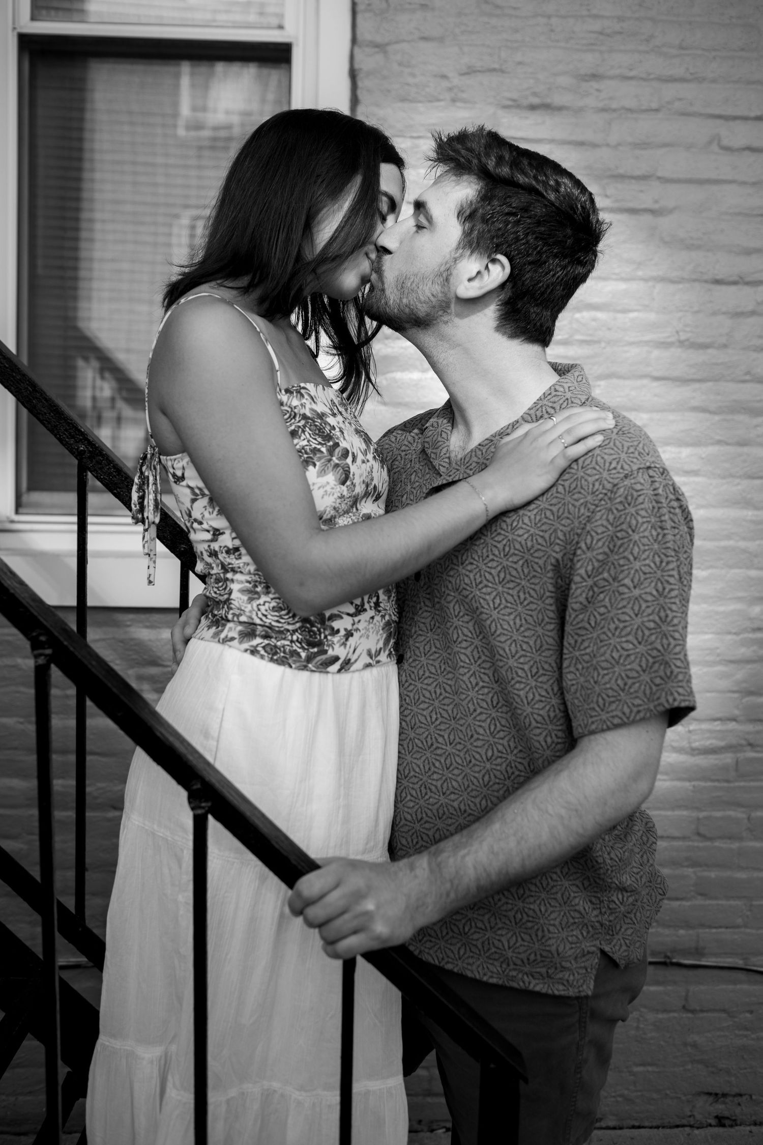 Black and white photo of couple sharing intimate moment on staircase against white brick wall.