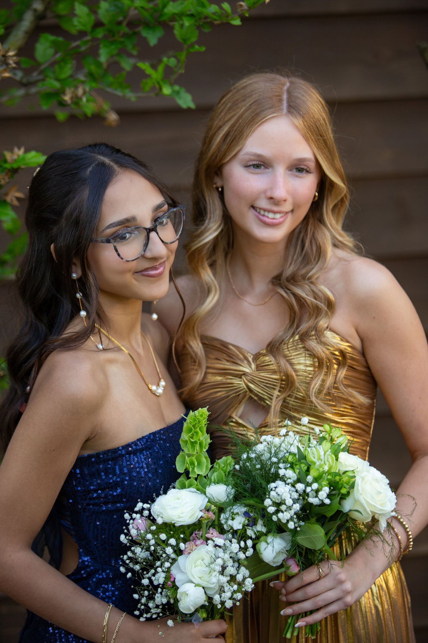 Two prom attendees wearing blue sequin and gold formal dresses pose together with a white flower bouquet.