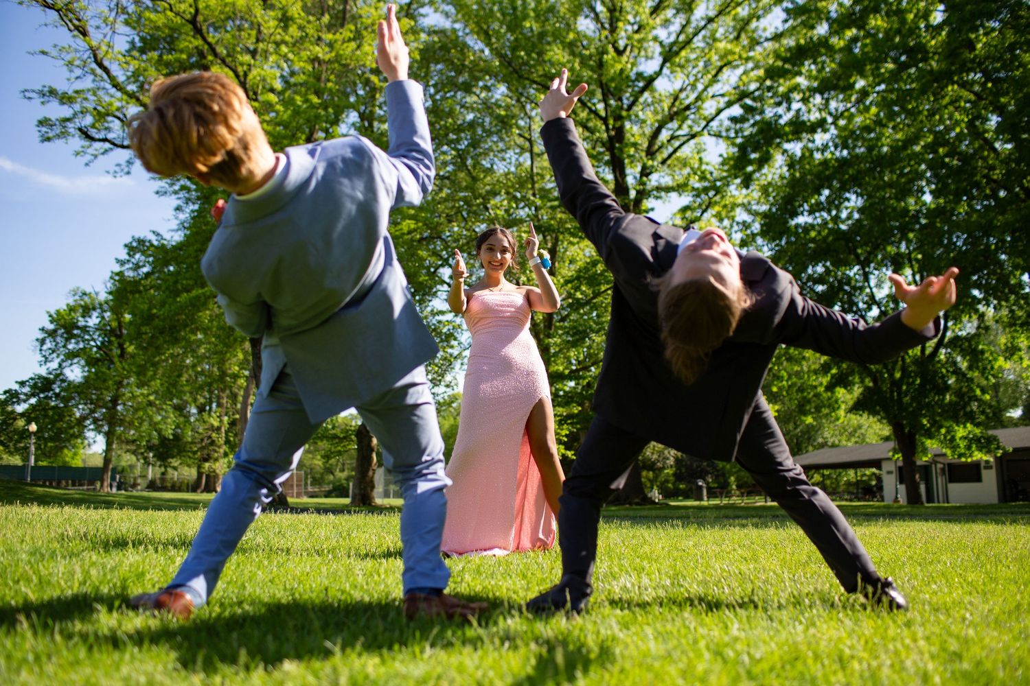 Three people strike dynamic dance poses on a sunny green lawn surrounded by trees during an outdoor performance.