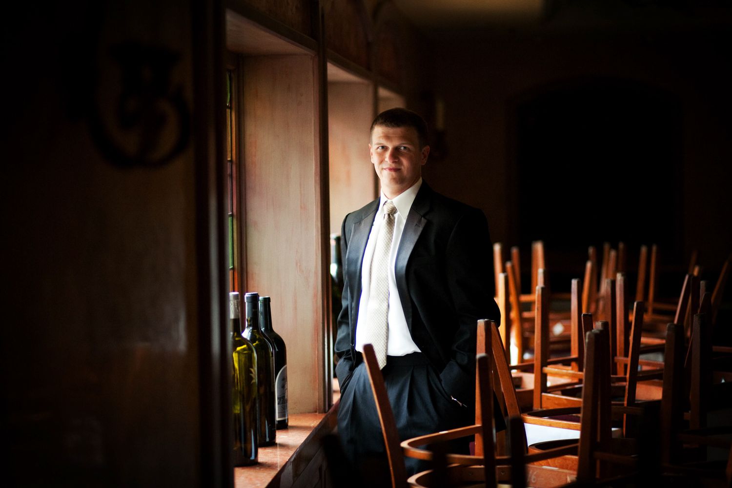 Man in formal attire stands contemplatively in a dimly lit room with stacked chairs.