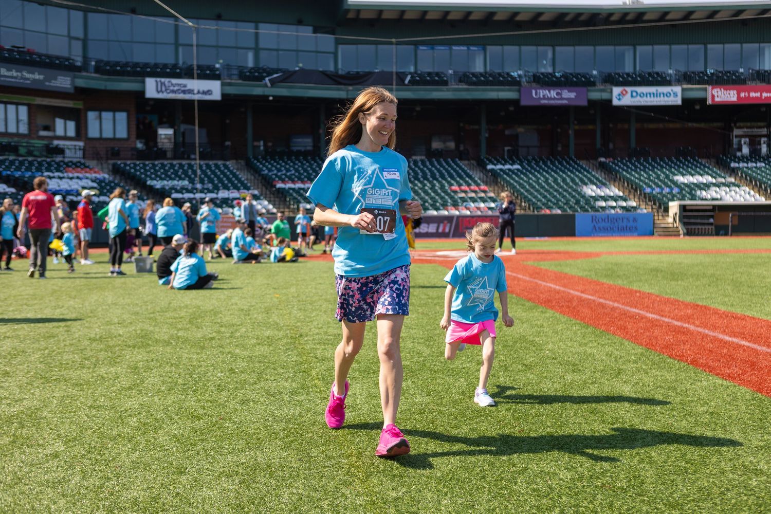 Participants in light blue shirts gather on a baseball field during a sunny outdoor event.