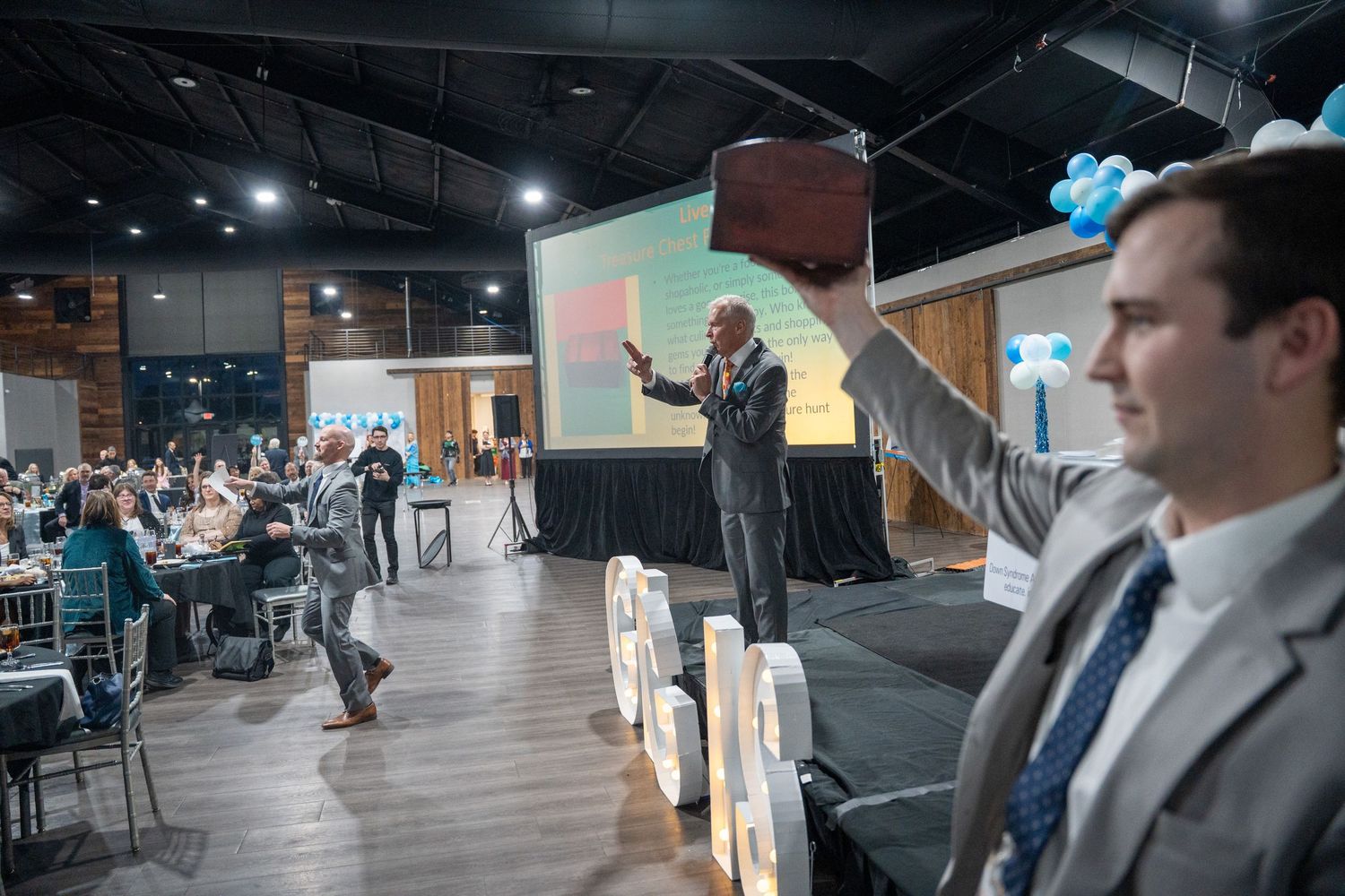 A speaker presents to an audience in a modern conference room with wood floors and cafe-style seating area.