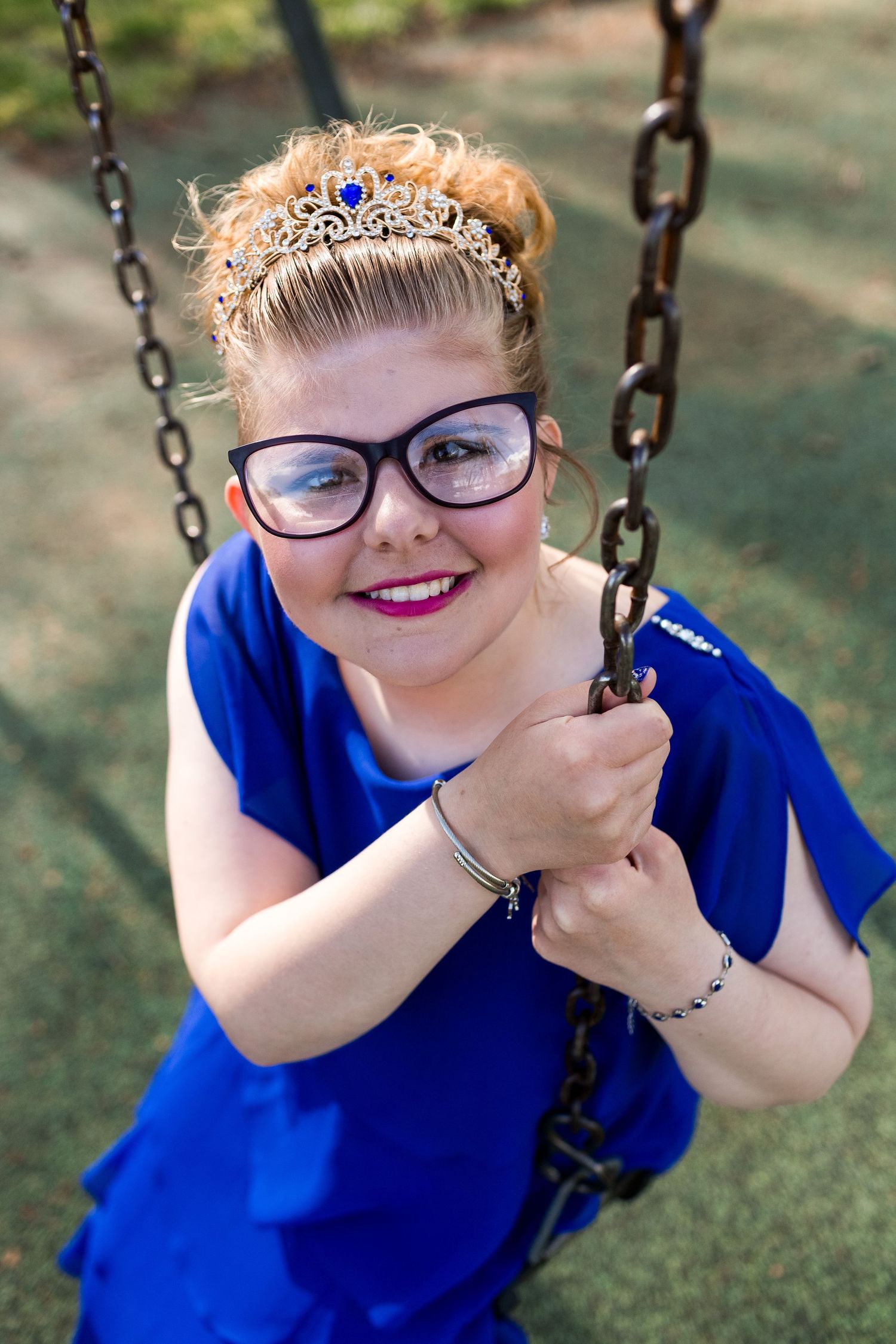 Someone wearing glasses and a blue dress sits on a swing outdoors while wearing a decorative crown and smiling warmly.
