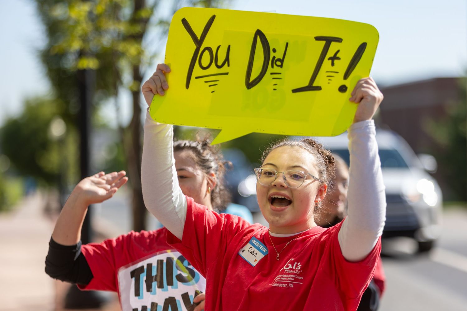 Young person holding yellow 'You Did It!' sign with enthusiasm at outdoor event.