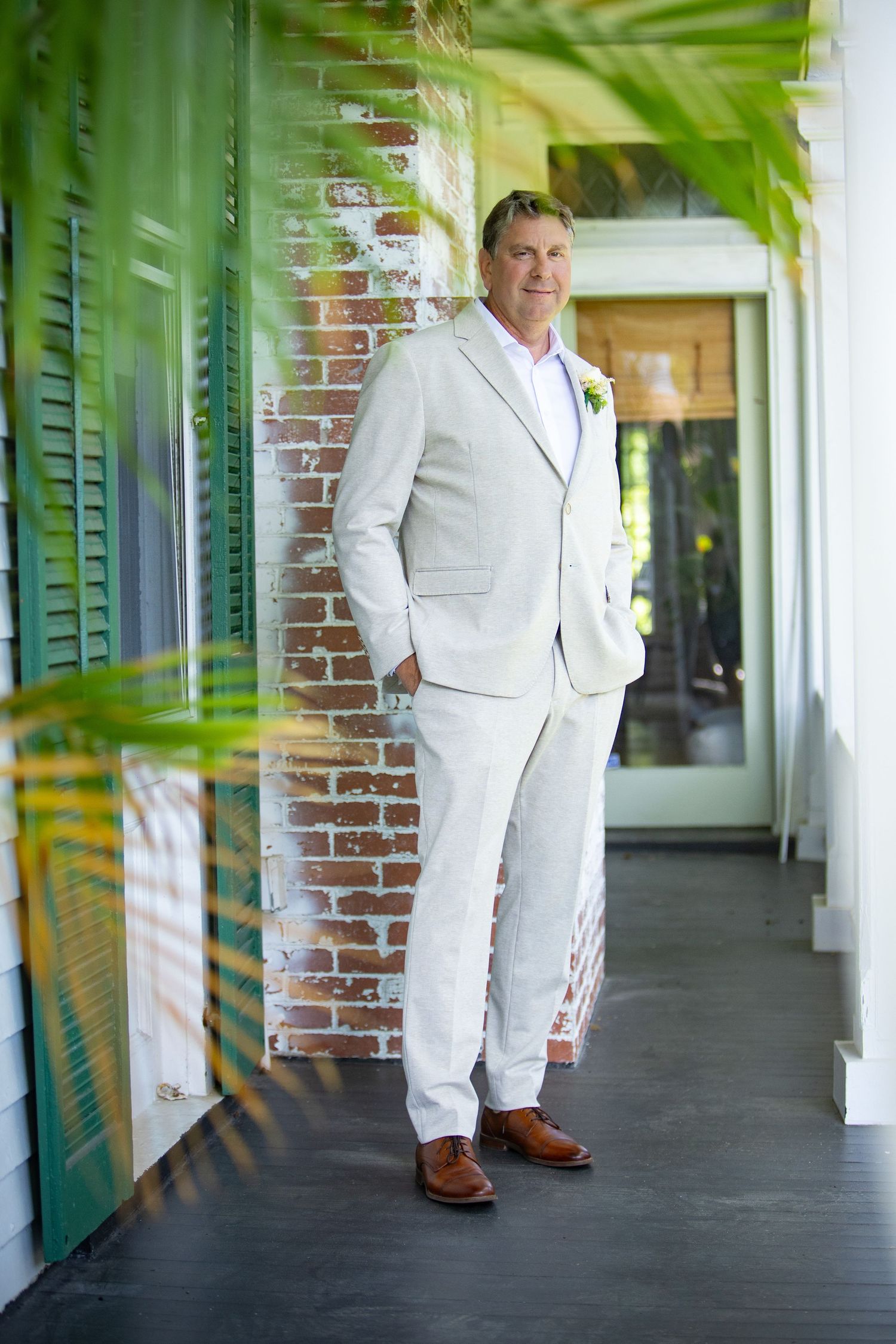 A groom in a light colored suit stands on a covered porch with brick walls and tropical decor.