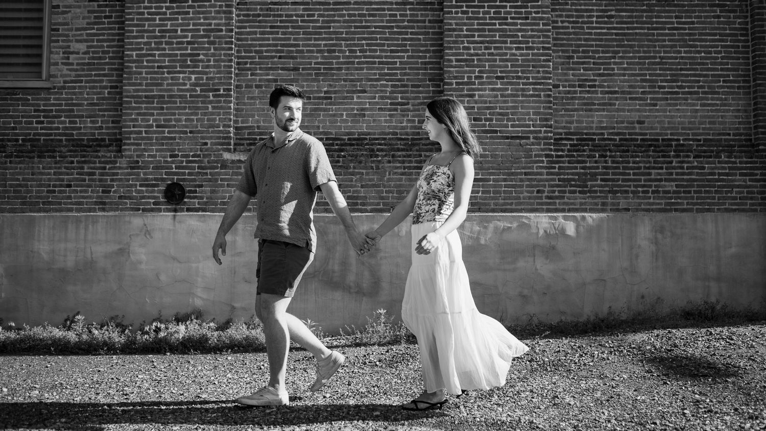 Black and white photo of couple in formal attire playfully running together in front of brick wall.