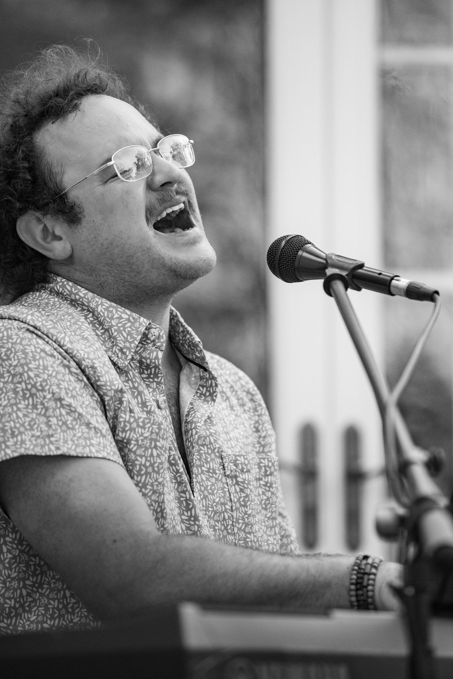 A musician in glasses plays piano and sings joyfully into a microphone at an outdoor performance.
