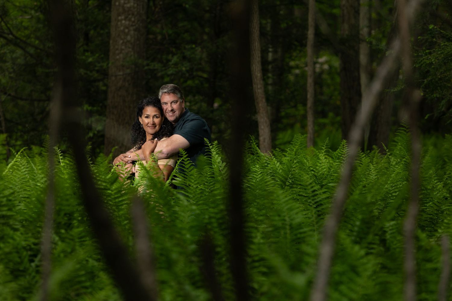 Two people sit together surrounded by lush green ferns in a moody forest setting.