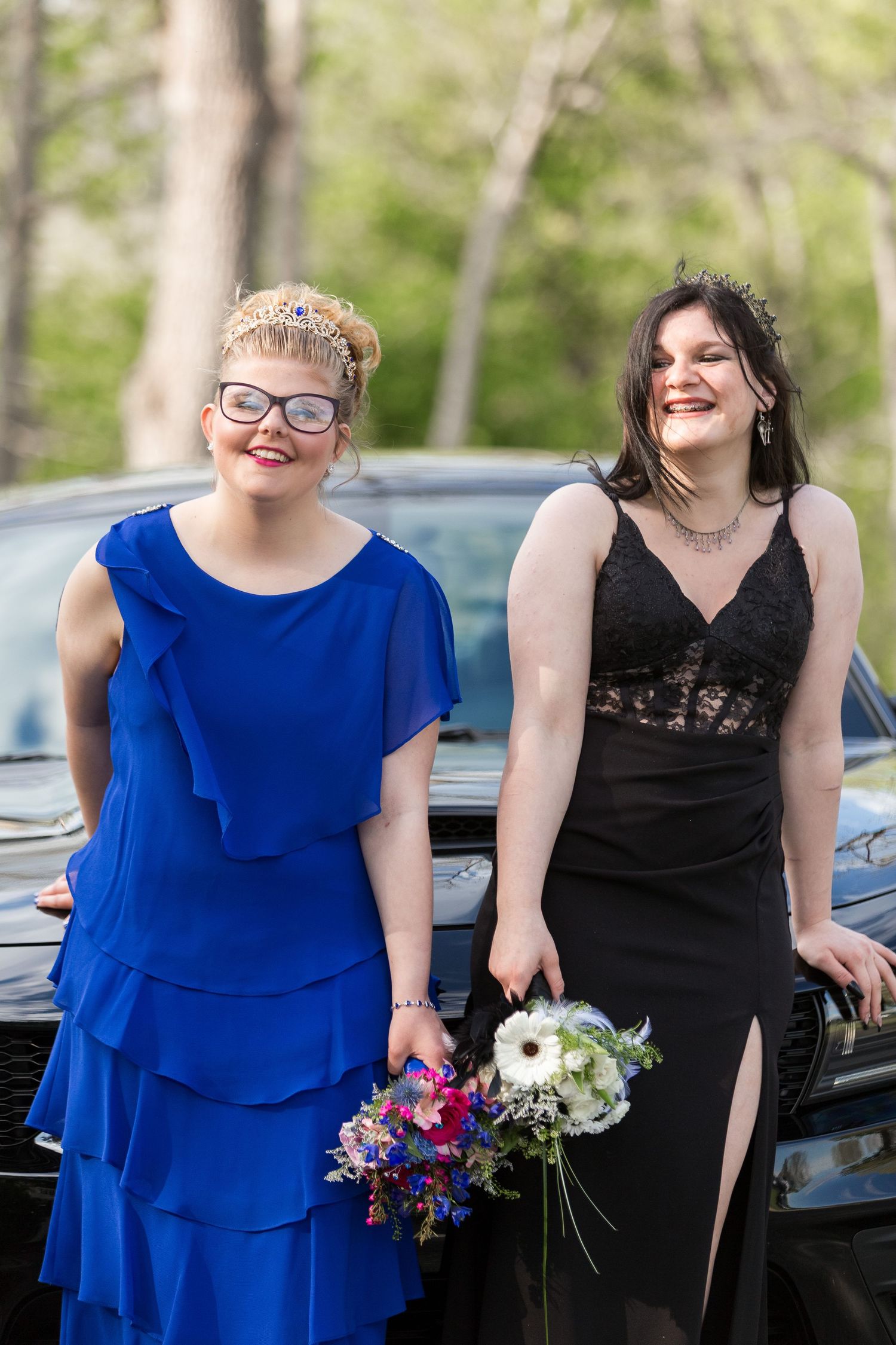 Two people in formal dresses lean against a car holding flower bouquets at a special event.
