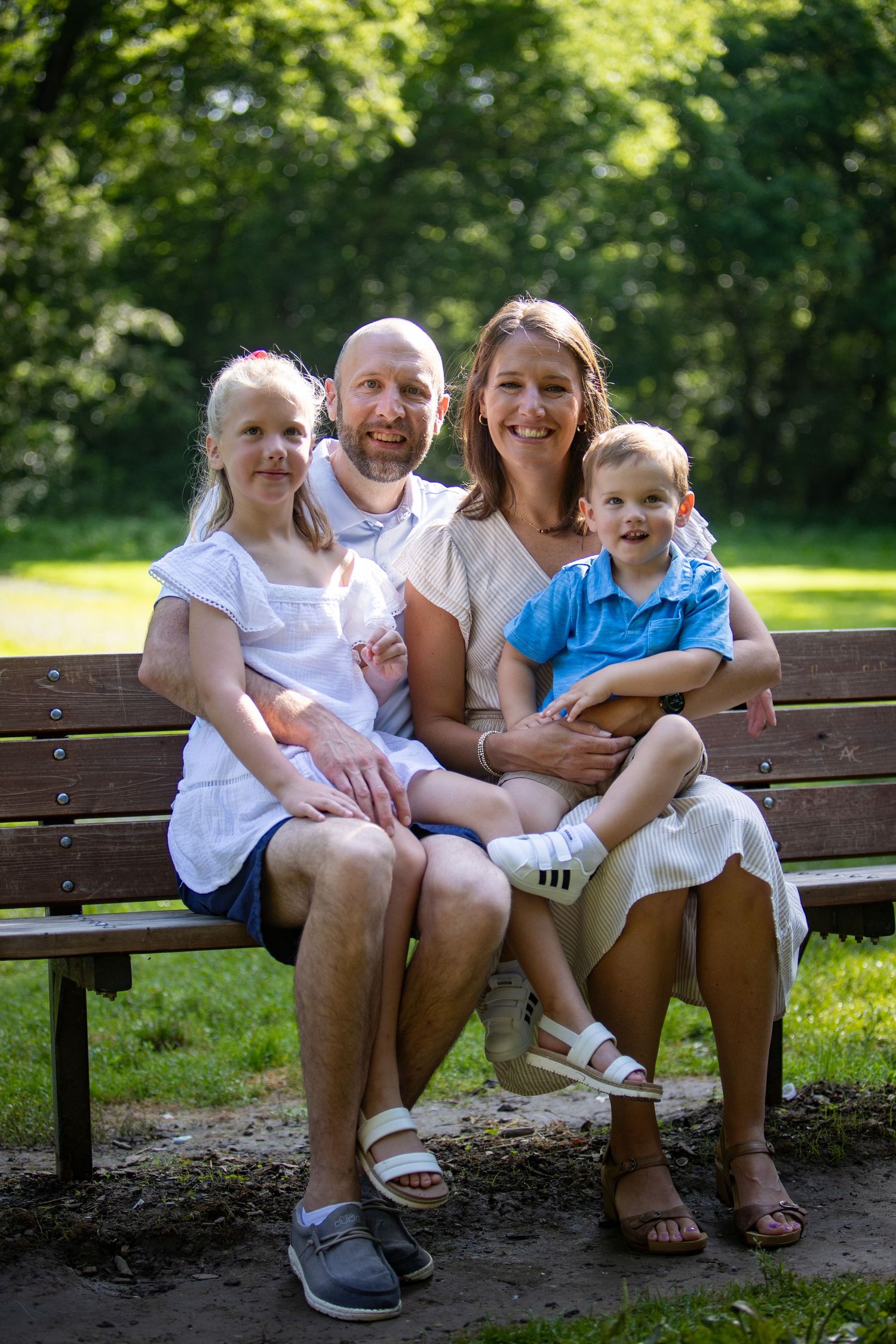 A happy family sitting together on a wooden park bench in a lush green outdoor setting on a sunny day.