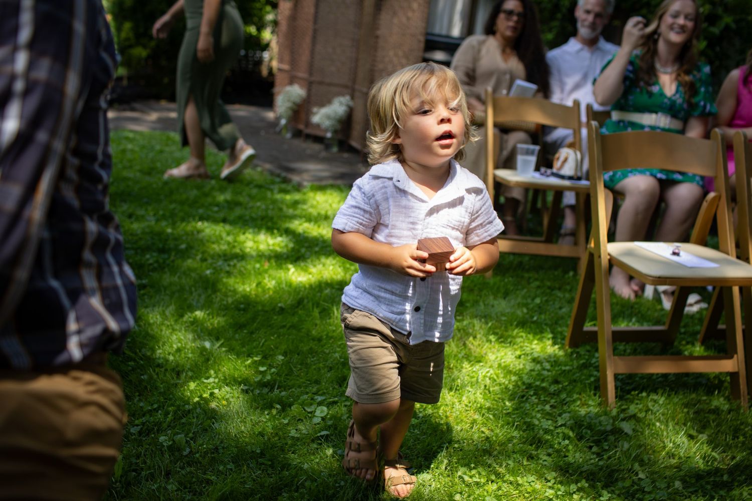 Child in striped shirt and khaki shorts playing on sunny grass lawn with folding chairs in background.