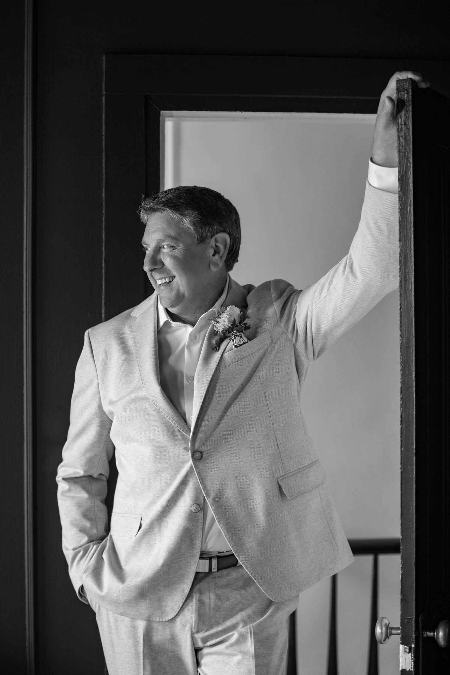 A groom in a light colored suit stands confidently in a doorway in black and white.