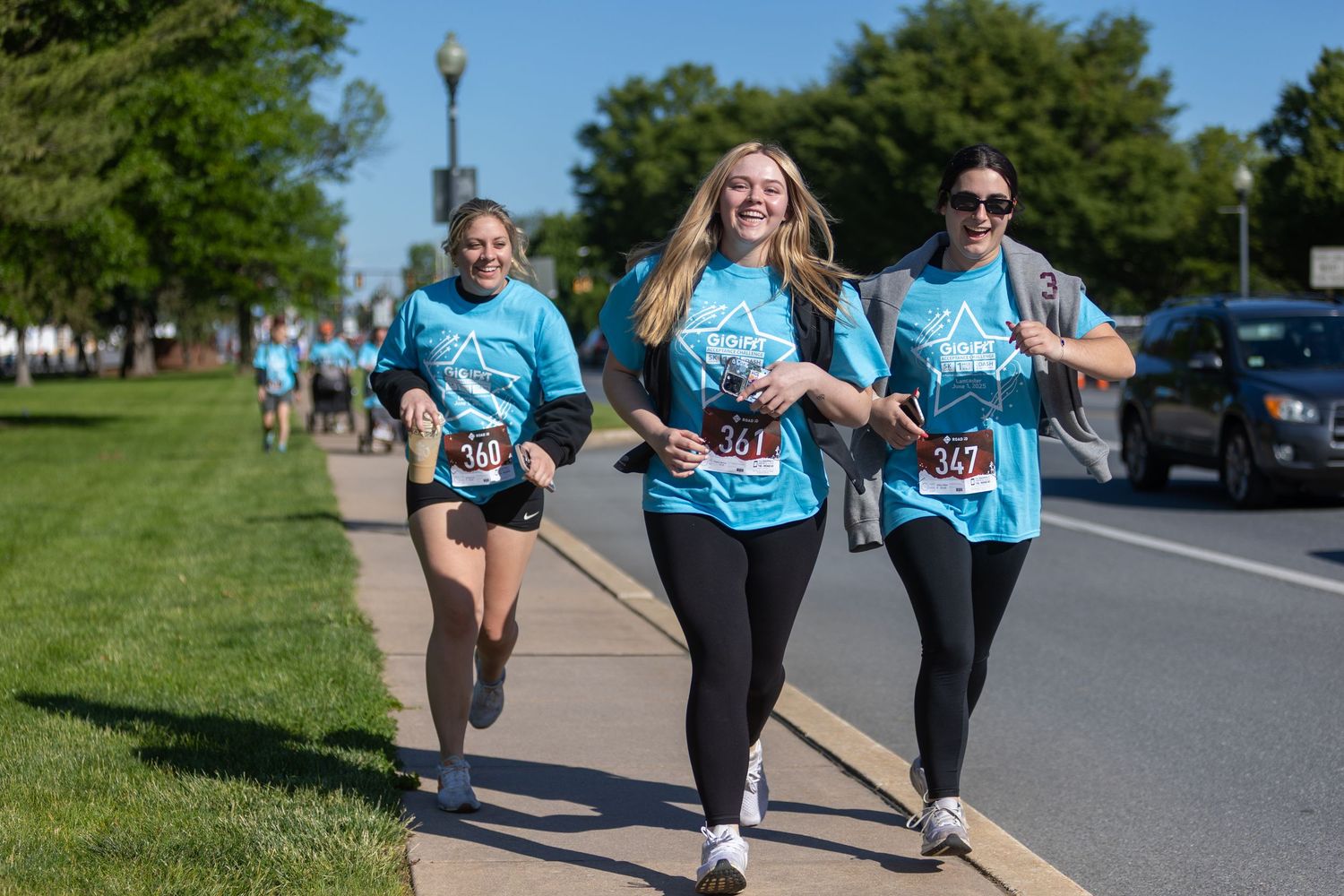 Group of runners in matching light blue shirts jog along a sidewalk during a community event.