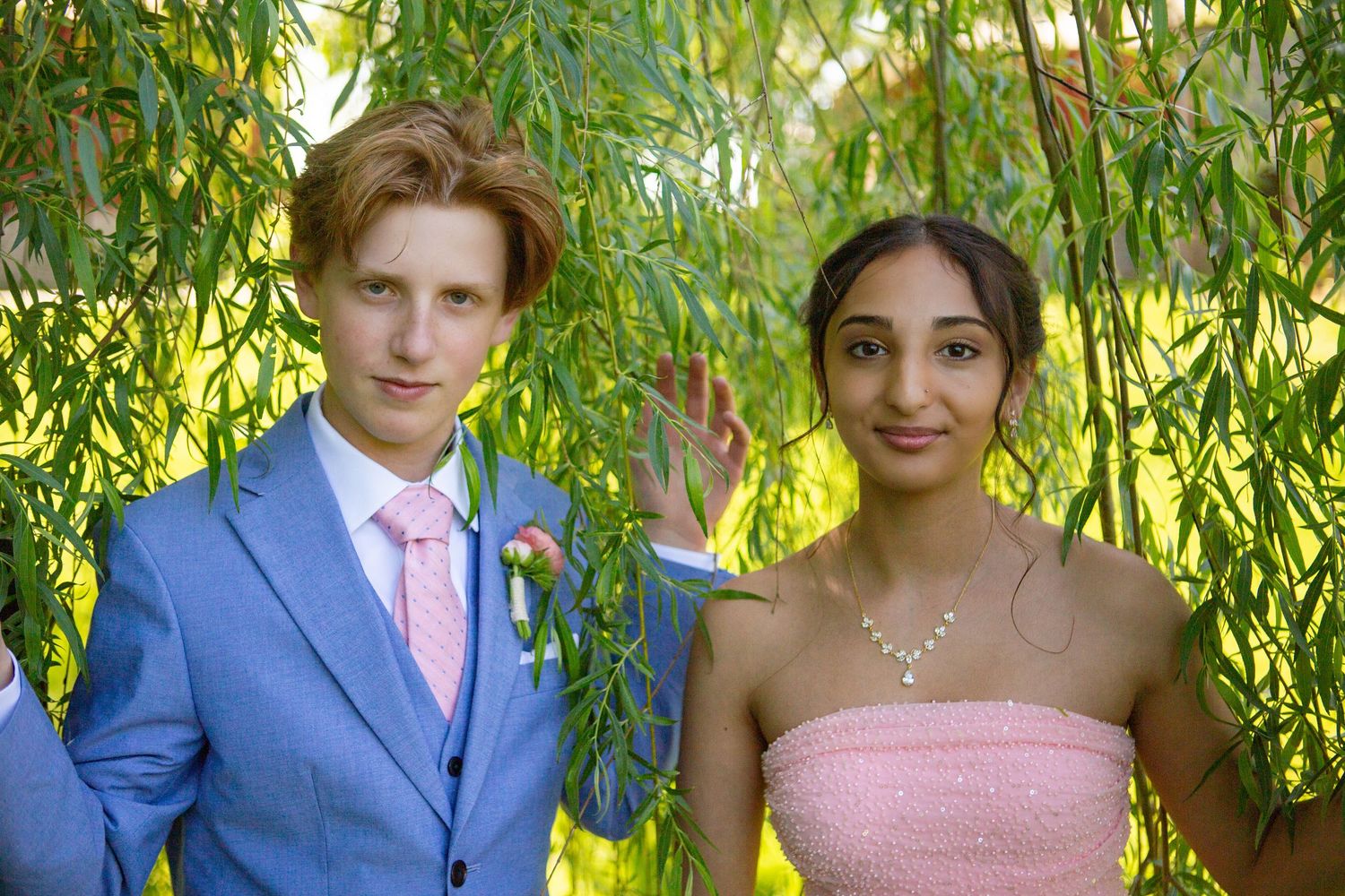 Two wedding guests in coordinating blue and pink formal attire pose together under graceful willow tree branches.