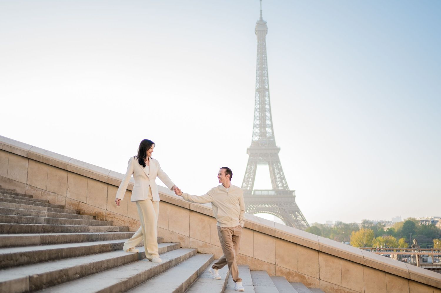 A couple in coordinating beige outfits walks down the Trocadéro steps with the Eiffel Tower visible in the background.
