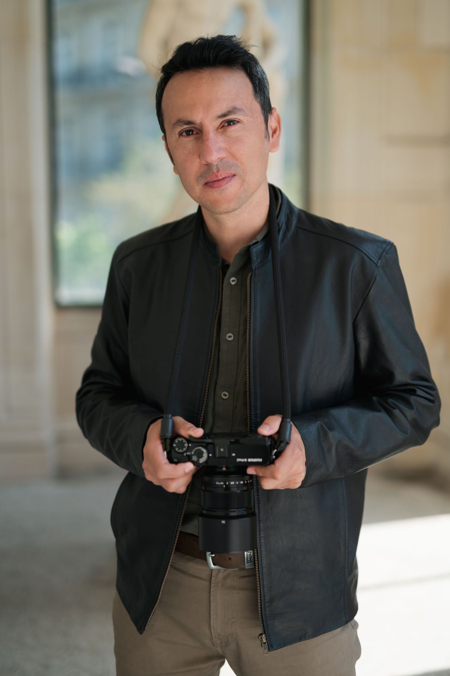 Professional photographer holding a DSLR camera while wearing a black leather jacket and khaki pants.