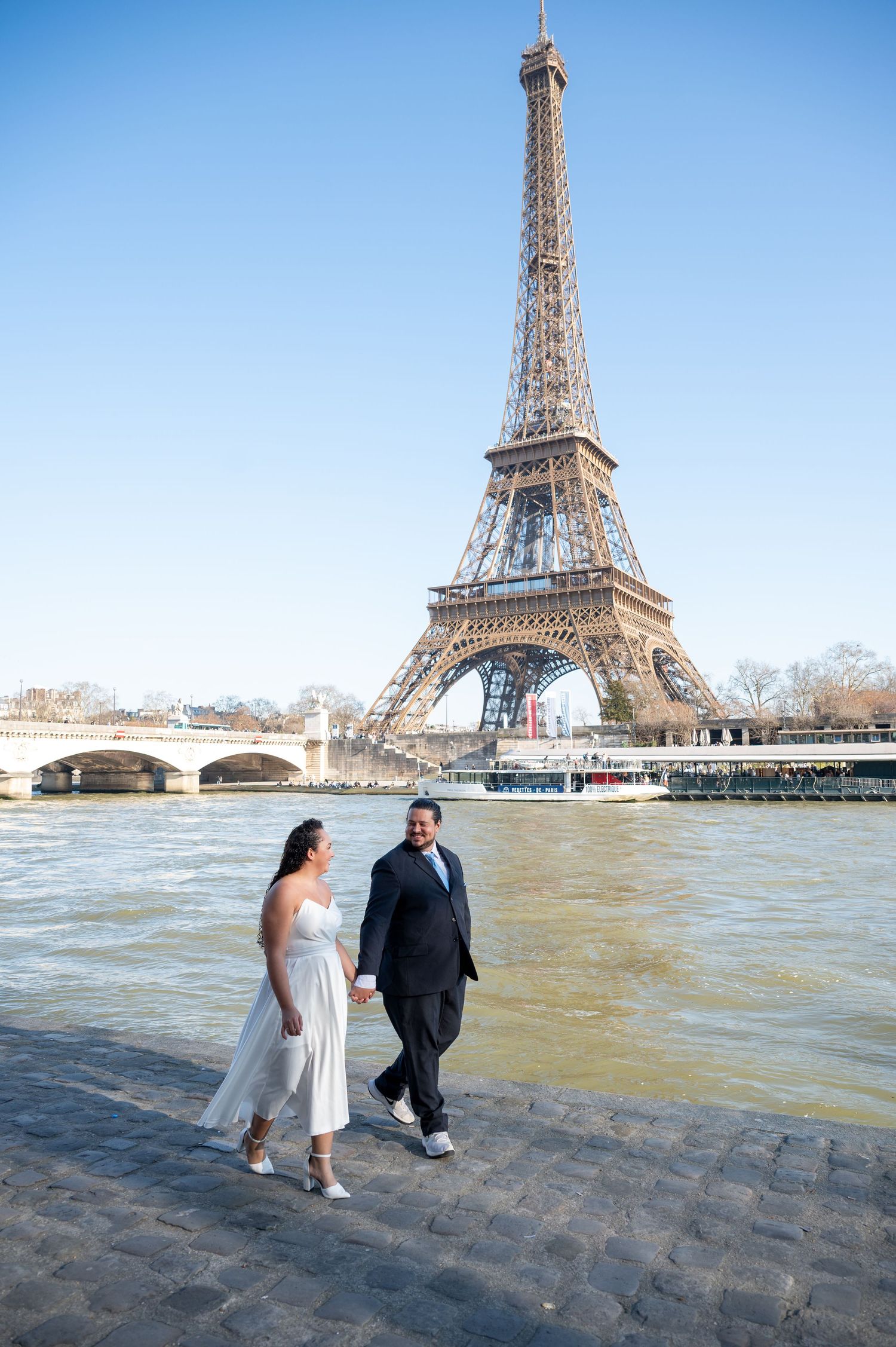 A couple strolls along the cobblestone riverbank of the Seine with the iconic Eiffel Tower rising majestically in Paris.