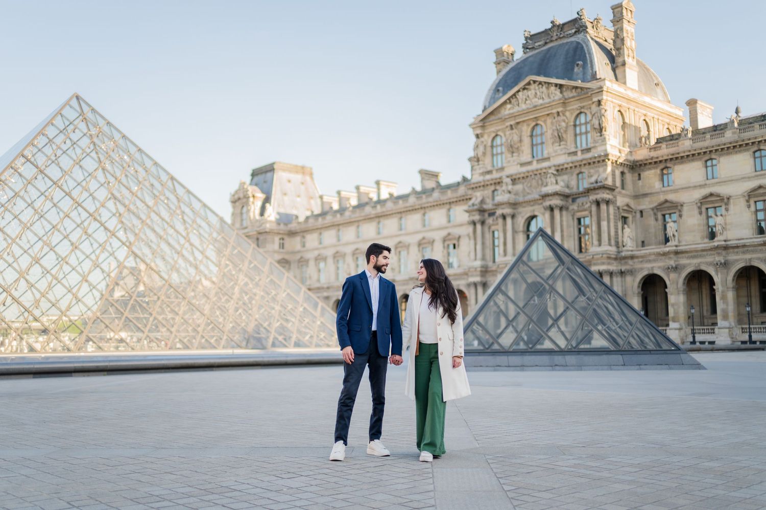 A couple strolls in front of the iconic glass pyramid at the Louvre Museum in Paris during a beautiful morning.