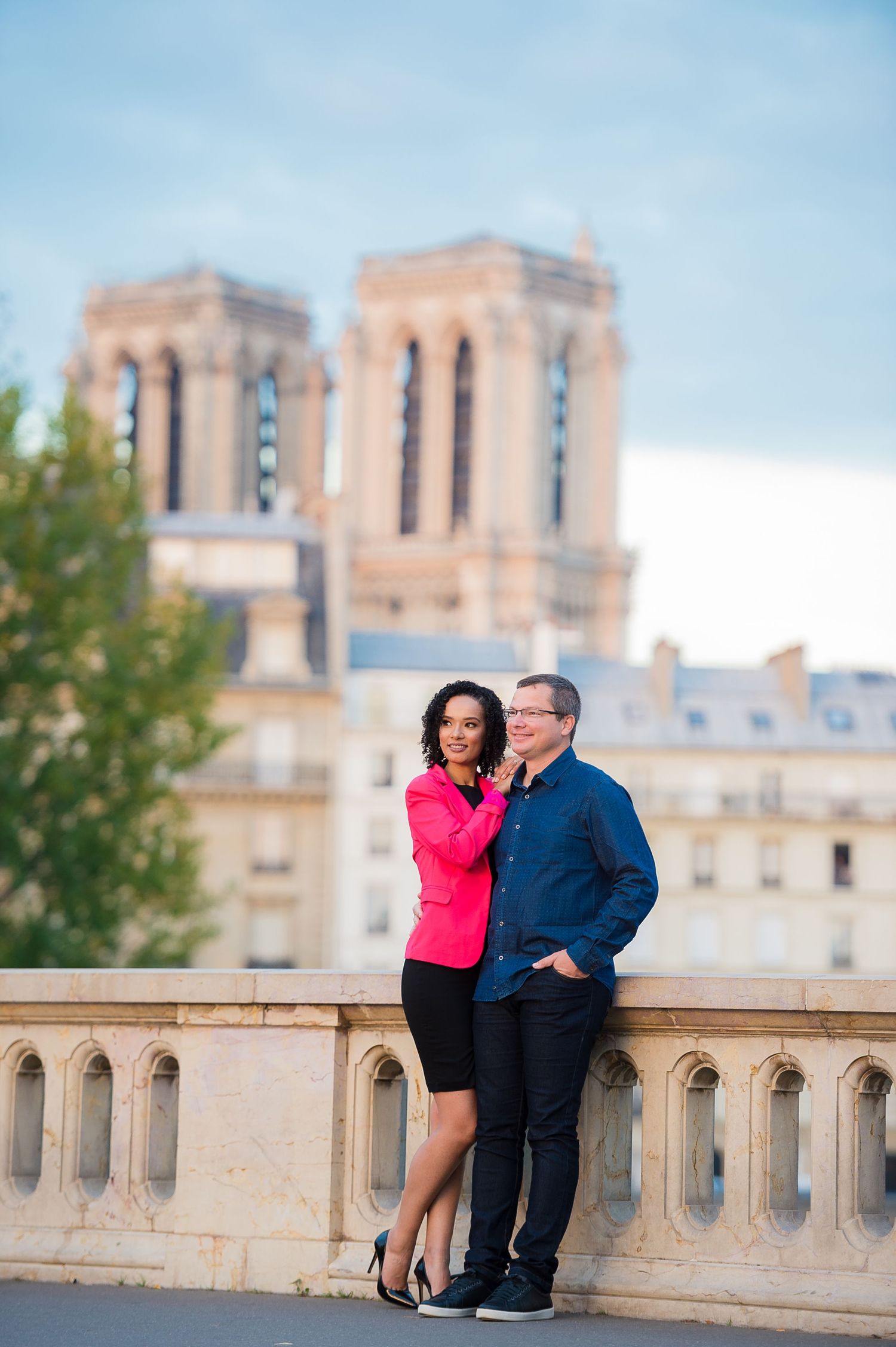 A couple poses romantically on a bridge near Notre Dame Cathedral in Paris with its iconic towers visible in the background.