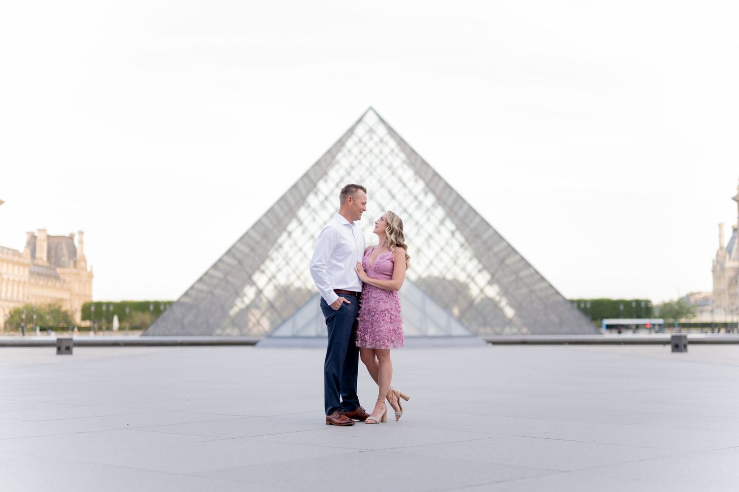A couple in romantic pose stands in front of the iconic glass pyramid at the Louvre Museum in Paris.