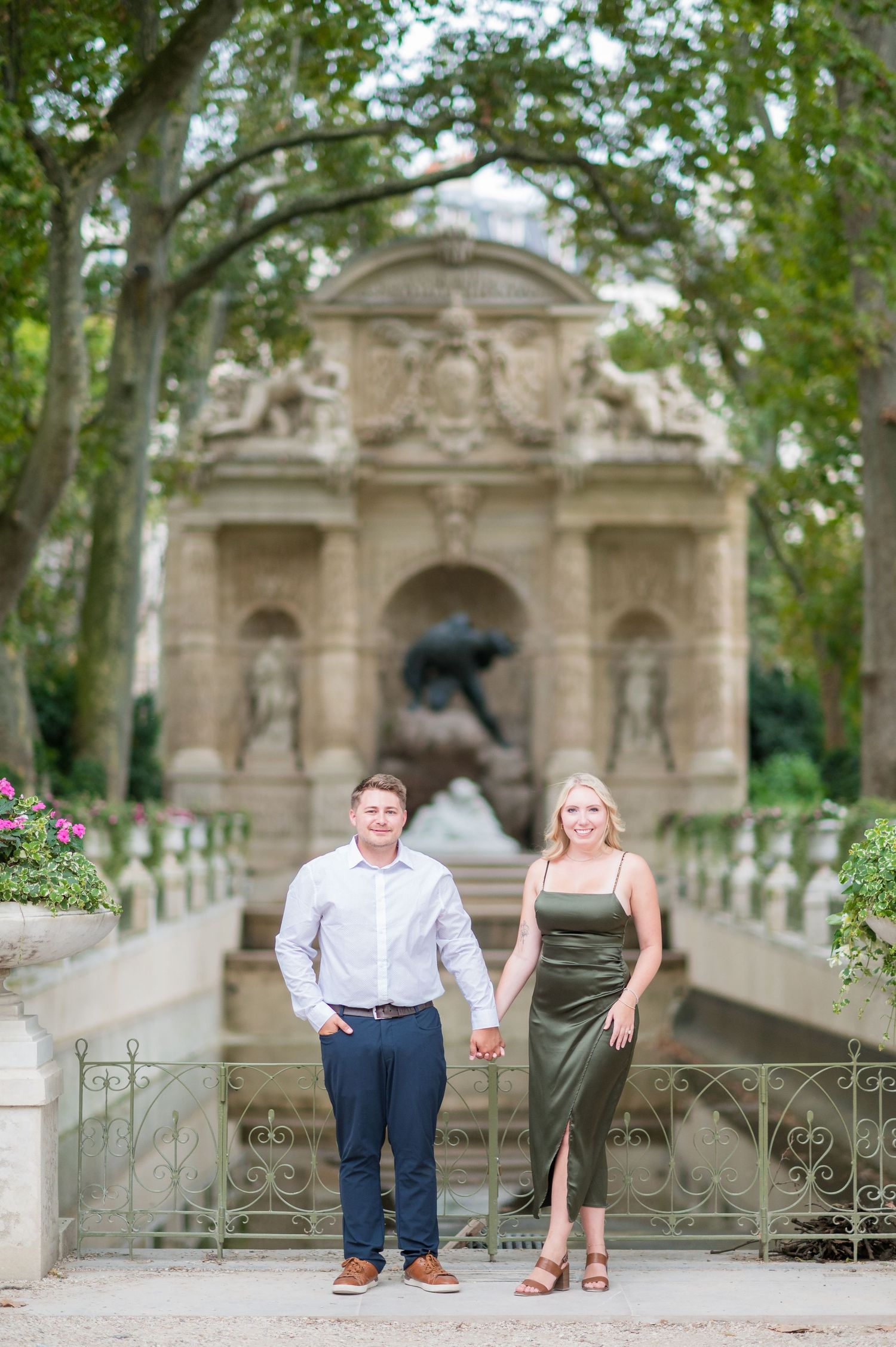 A couple holds hands in front of an ornate Parisian fountain surrounded by trees.
